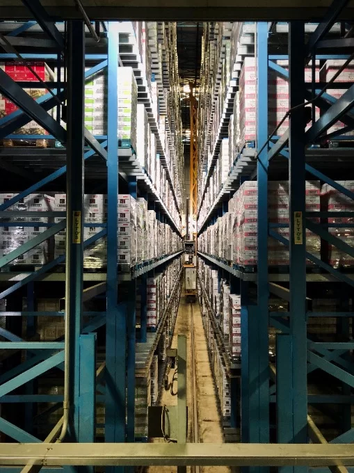 Perspective view down a narrow aisle between tall shelving stocked with boxes in a large warehouse.